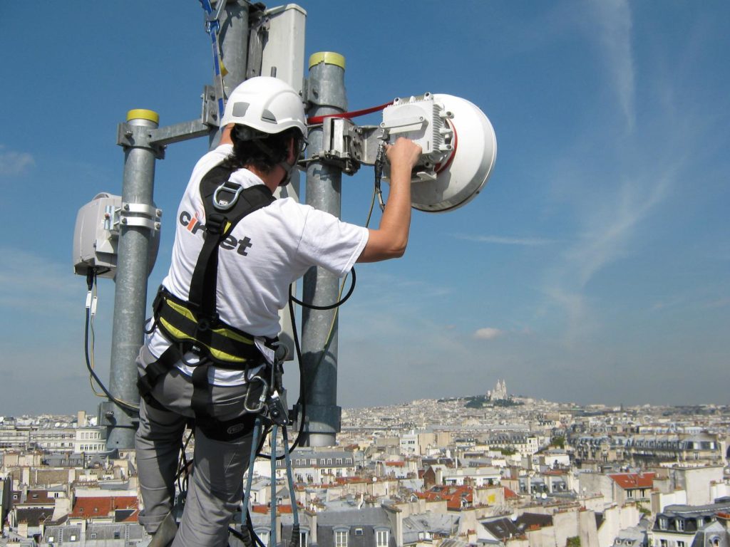 A telecommunication engineer works on a cell tower in the foreground with the Paris skyline in the background