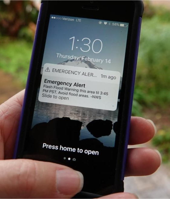 A hand holding a mobile phone with an Emergency Alert notification for a flash flood warning on the home screen